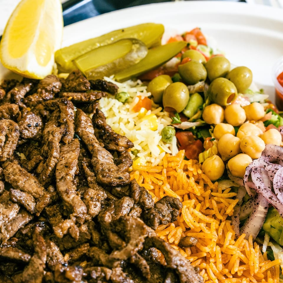 Close-up of a Casa De Falafel plate with beef shawarma, rice, chickpeas, olives, pickles, and lemon.